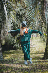 A woman in an emerald corduroy suit with a hat covering her face stands on a lawn among the palm trees. Imitation of a garden scarecrow