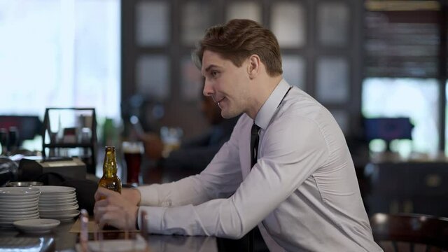 Side view of relaxed businessman drinking beer ordering more alcohol at bar counter. Portrait of successful young handsome Caucasian man enjoying Friday. Blurred African American guy at background