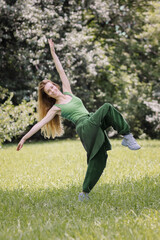 A woman in green clothes in a pose on one leg spreading her arms to the sides stands in the park on...