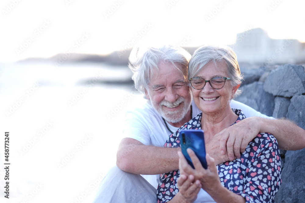 Wall mural smiling senior couple embracing in outdoor at sea at sunset using mobile phone. caucasian retired en