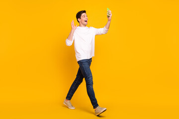Full body photo of cute brunet young guy go talk telephone wear shirt jeans sneakers isolated on yellow background