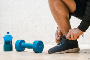 man tying his shoes next to dumbbell and bottle with blue water, before exercising. exercise at home, white background. fitness concept. health concept, space for advertising text