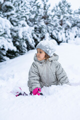 Fototapeta na wymiar Kid Sits in a snowdrift near the Christmas trees. Winter games for children with snow. Happy child in a snowdrift