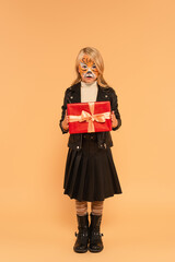 full length view of child in stylish clothes and tiger makeup holding present on beige