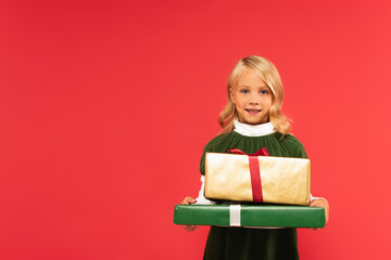 cheerful girl smiling at camera while holding presents isolated on red