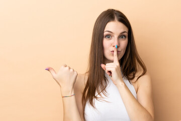 Young caucasian woman isolated on beige background pointing to the side and doing silence gesture