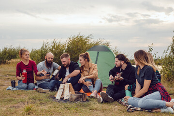 A group of friends sit by the fire on the background of the river and sing to the guitar.