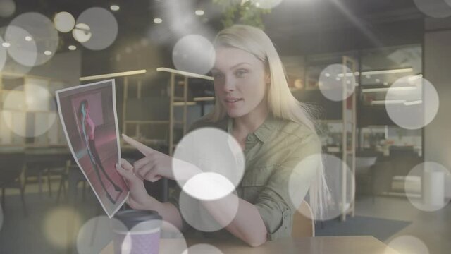 Animation of light spots over caucasian businesswoman having video call and showing photos