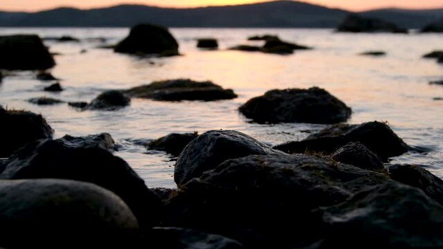 slow motion, a warm summer evening sunset on the shore of the mora through the strait, the sun almost went down behind the mountains, there are many stones on the near shore