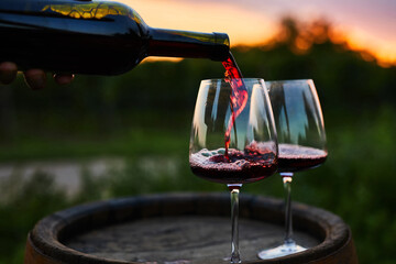 Pouring red wine into glasses on the barrel at dusk - obrazy, fototapety, plakaty