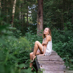 Naklejka premium Young caucasian natural woman in a white short dress in a summer forest. Girl with blond hair sits barefoot among the green trees on a wooden bridge and smiles