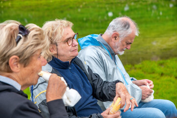 Elderly people relaxing at the end of a mountain hike, eating sandwiches on the grass.