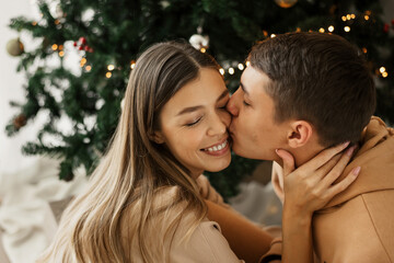 Fototapeta premium Young couple sitting near Christmas tree at home. Man kissing a woman. Love and relationships. Christmas and New Year celebration