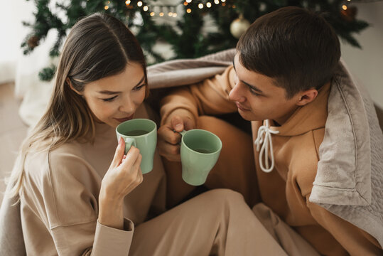 Young Couple Wearing Comfy Loungewear Drinking Tea Near Christmas Tree. Christmas Morning. Winter Holidays