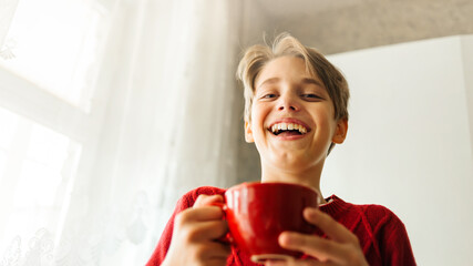 Happy laughing teenager boy holds a red cup with cocoa and marshmallows. Soft selective focus. © Viktoriia