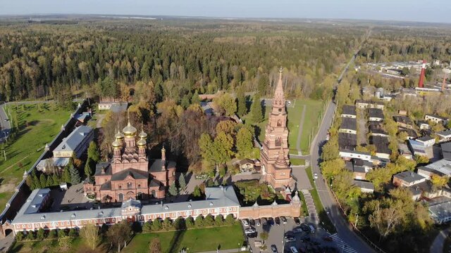 Aerial drone view of bell tower and red-brick cathedral on the background of the river at sunny autumn day. Gethsemane Chernigov Skete, Sergiyev Posad, Russia in 4K