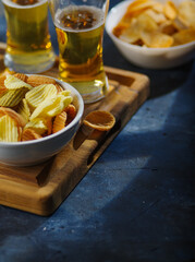 Amber beer in glasses and a bowl of potato chips on a wooden tray. Low angle view. Blue background. Rest, relaxation in the company of friends, watching TV and sports matches.