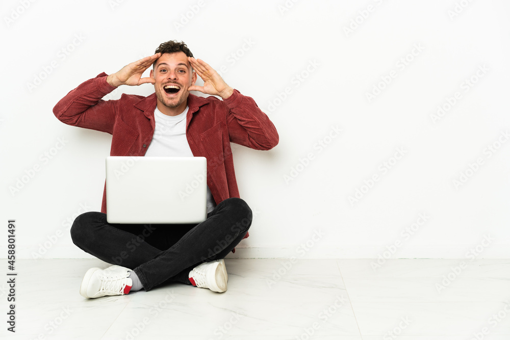 Wall mural Young handsome caucasian man sit-in on the floor with laptop with surprise expression