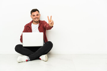 Young handsome caucasian man sit-in on the floor with laptop smiling and showing victory sign