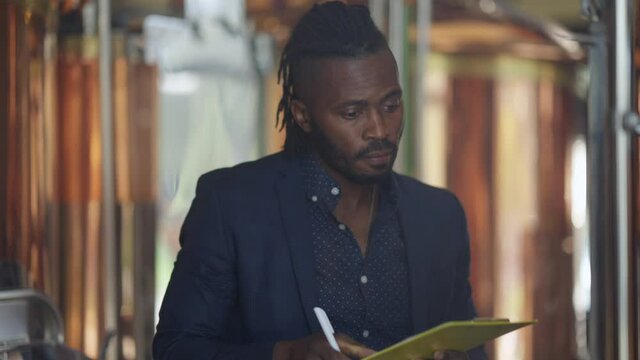 Expert African American brewer checking beer quality filling in paperwork standing at large tanks indoors. Handsome concentrated professional man working at brewing factory