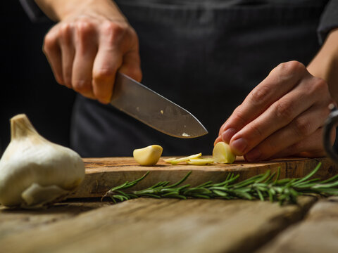 The chef cuts the garlic on a cutting board. A sprig of rosemary. Wooden texture. Macro photography. Hot spice for salads, sauces, soups, meat, fillings. Restaurant, hotel, cafe, home cooking.