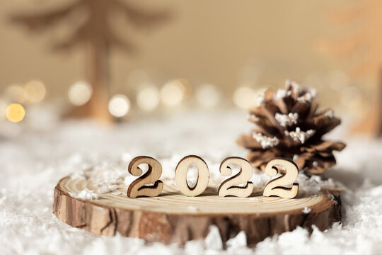 New year 2022. Numbers 2022 on wooden stand on beige pastel blurred background with decorative fir trees, cone, snow and lights. Christmas greeting card.