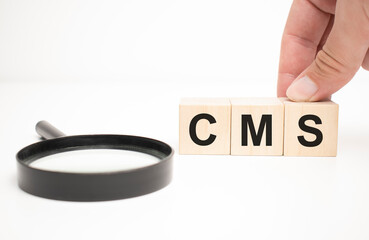 cms text wooden cube blocks and hand holding magnifying glass on table background.