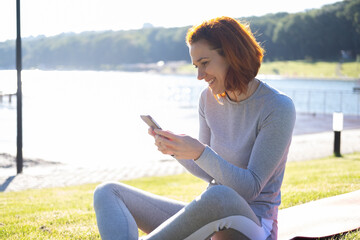 Naklejka premium slim ginger woman in sport clothes using mobile phone at park. Health, wellness, fitness lifestyle