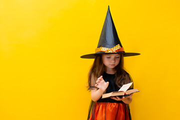 Halloween. A serious cute girl in a witch costume holds an old book in her hand. Colored Yellow background. A place for text.