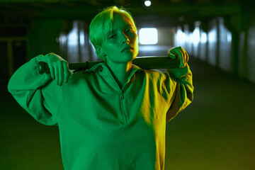 a daring girl with a bat in her hands stands in an empty parking lot and looks directly into the camera. Modern shooting in bright green light. High quality photo