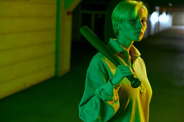 a teenage girl with short hair holding a baseball bat and looking straight into the camera. Modern parking in neon green light. High quality photo