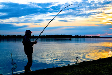 A fisherman with a spinning pulls the caught fish out of the water. Silhouette at sunset.