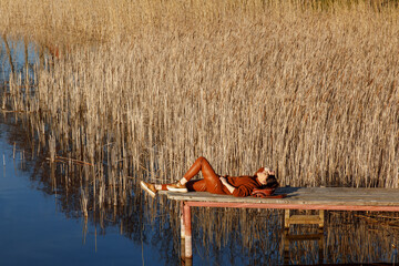 Portrait of a woman in autumn cozy orange clothes with backpack sitting on a wooden bridge near the lake with blue water and looking agains the sun. People, travel, nature concept.