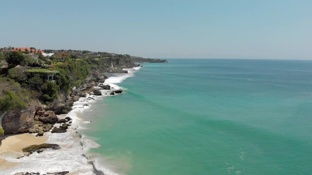 Alex Drone shot of magnificent seashore. Blue waves breaking on rock coast where hoses placed inbetween trees. Tropical seascape on bright day shot by drone. Concept of travel, exotic, landscape.