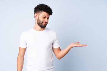 Young Moroccan man isolated on blue background holding copyspace with doubts