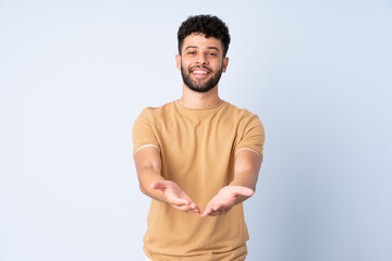 Young Moroccan man isolated on blue background holding copyspace imaginary on the palm to insert an ad