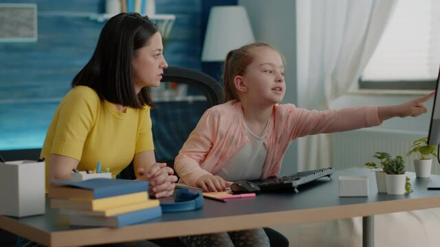 Child receiving assistance for online task from mother on computer at home. Parent helping little girl with homework and remote school lessons, pointing at monitor. Schoolgirl and mom
