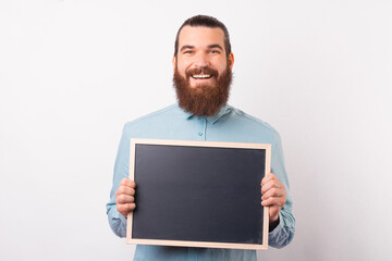 Smiling bearded man is holding a black chalkboard in front of him over white background.