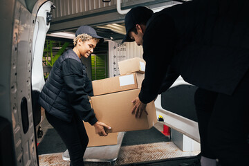 Two couriers stack cardboard boxes in a delivery van. Coworkers pick up the packages from the...