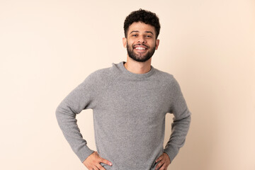 Young Moroccan man isolated on beige background posing with arms at hip and smiling