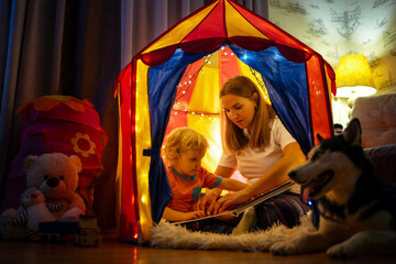 Obraz na płótnie Canvas Smiling little boy with mother are reading a book in tent with garlands at home. Concept of child education and family having time together at night