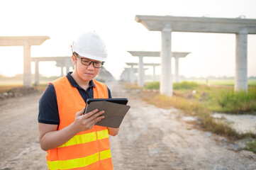 Fototapeta premium Smart Asian worker man or male civil engineer with protective safety helmet and reflective vest using digital tablet for project planning and checking architectural drawing at construction site.