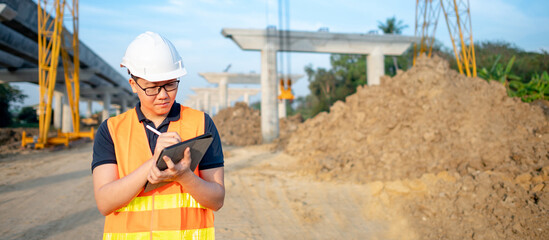 Smart Asian worker man or male civil engineer with protective safety helmet and reflective vest using digital tablet for project planning and checking architectural drawing at construction site.