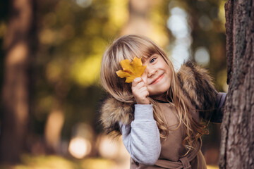 Beautiful blonde girl covers one eye with yellow maple leaf outdoors in autumn park.