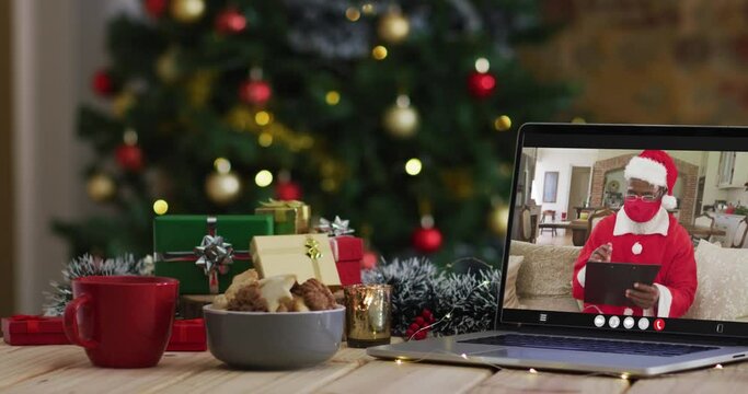 African american santa in face mask on video call on laptop, with christmas decorations and tree