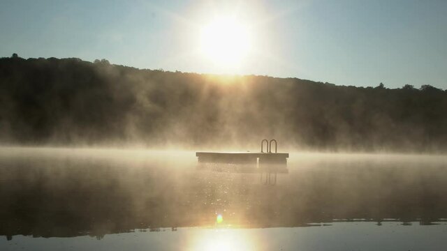 A swim dock is seen through the mist in the early morning sunrise.
