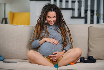 Joyful pregnant woman with dreadlocks sits on comfortable couch in living room and gentle touching...