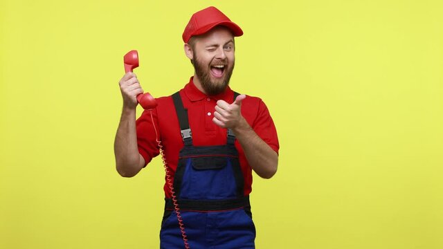 Positive handsome call centet worker talking via retro landline phone and showing thumb up, wearing blue uniform, red T-shirt and visor cap. Indoor studio shot isolated over yellow background.