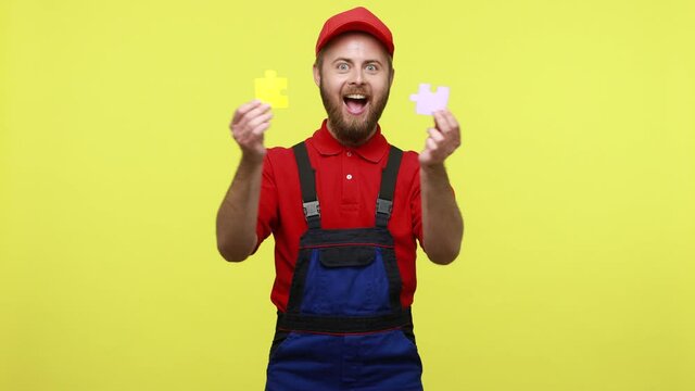 Happy smiling worker man connecting two pieces of puzzles, solving hard task, integration, wearing blue uniform, red T-shirt and visor cap. Indoor studio shot isolated over yellow background.
