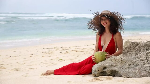 Alex Portrait of woman in red dress sitting on beach coast, holding coconut, serving as a container for drink. Girl in big straw hat, covered in white sand drinking coconut milk and smiling on camera.